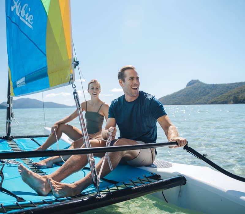 A man and woman sailing a blue catamaran on clear blue water near Hamilton Island.