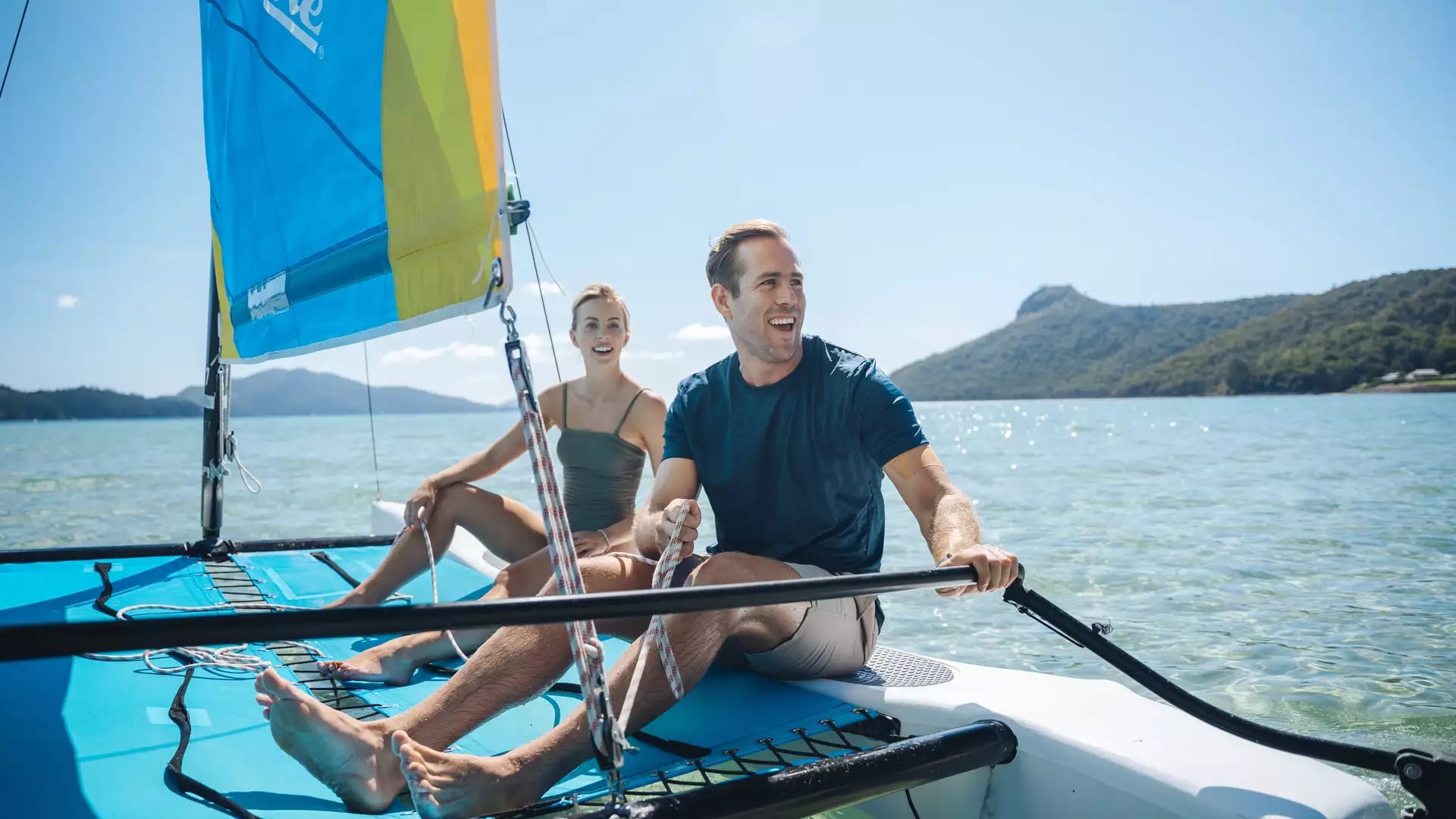 A man and woman sailing a blue catamaran on clear blue water near Hamilton Island.