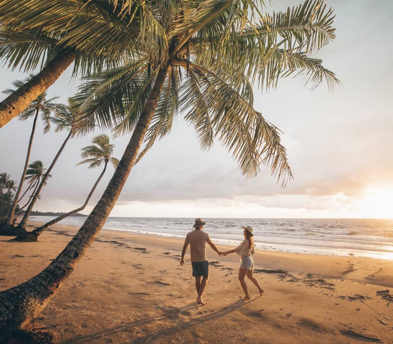 Silhouette of a couple walking on a beach at sunset framed by a large leaning coconut palm.