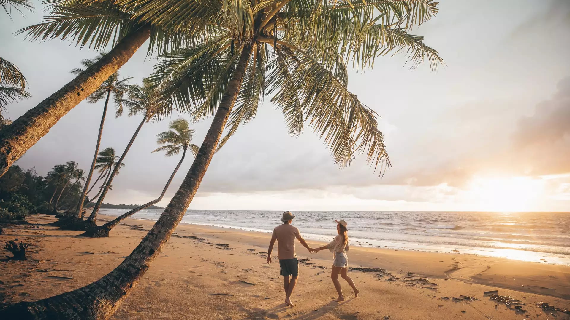 Silhouette of a couple walking on a beach at sunset framed by a large leaning coconut palm.