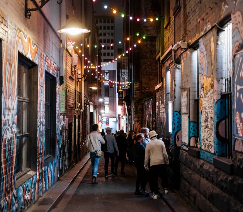 People walking through a famous Melbourne street art laneway at night under hanging string lights and vibrant murals.