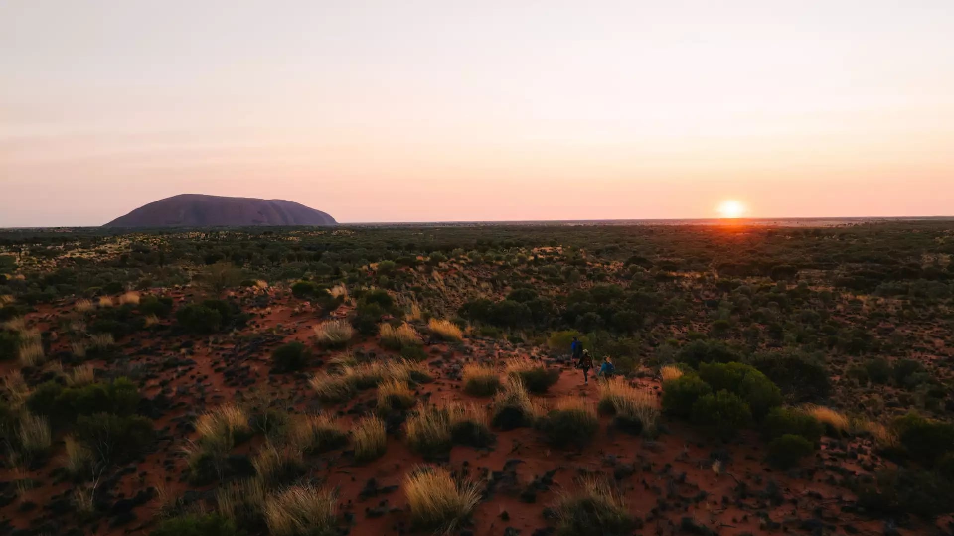 Wide landscape shot of hikers walking through desert scrub toward Uluru at sunrise in the Northern Territory.