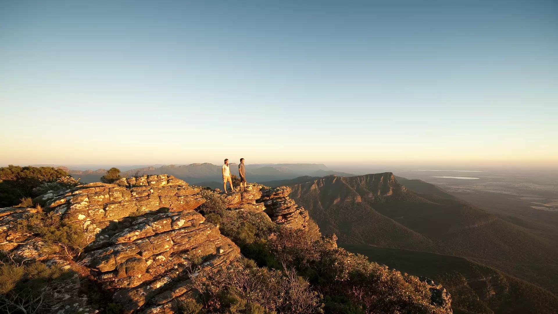 Two people stand atop a flat, weathered sandstone outcrop overlooking a vast, rolling forested valley and distant mountain ranges bathed in the soft golden light of a setting sun in The Grampians.