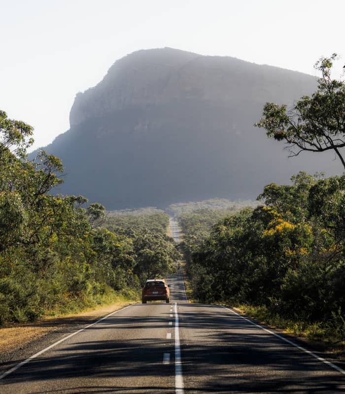 A brown SUV drives down a long, straight asphalt road that disappears into the distance toward a massive, hazy mountain peak surrounded by dense green bushland in The Grampians.
