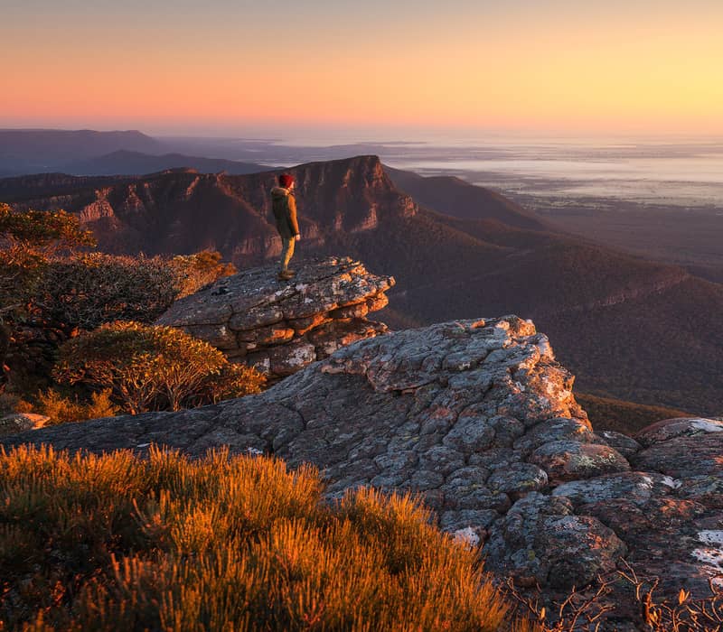 A hiker in a winter jacket stands on the edge of a rocky outcrop at The Pinnacle in The Grampians, looking out over a vast valley and distant mountain ranges under a glowing orange sunset sky.