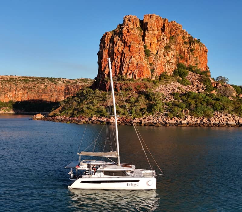 A high-angle aerial shot of the white catamaran Eclipse anchored in calm blue water. Behind it rises a massive, sheer orange-red sandstone cliff under a clear sky, with a small white tender boat moving nearby.