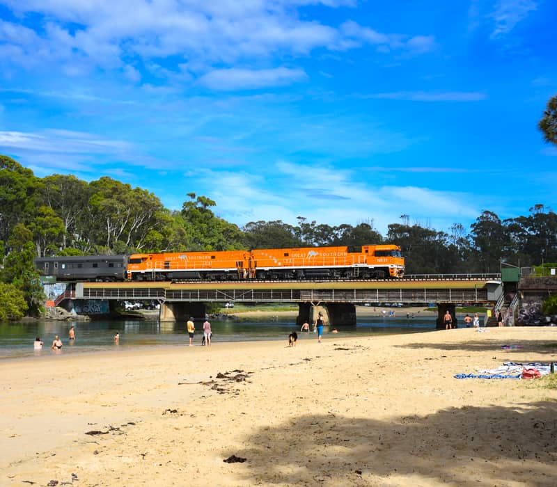 An orange passenger train crossing a bridge over a river with people on the beach in the foreground.