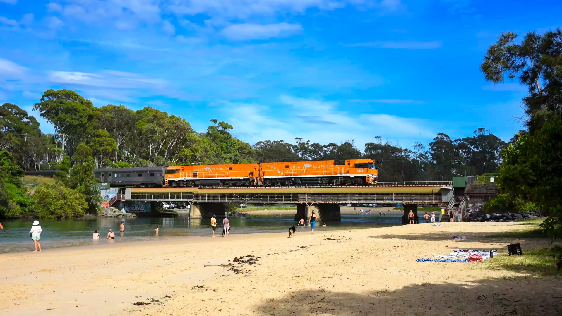 An orange passenger train crossing a bridge over a river with people on the beach in the foreground.