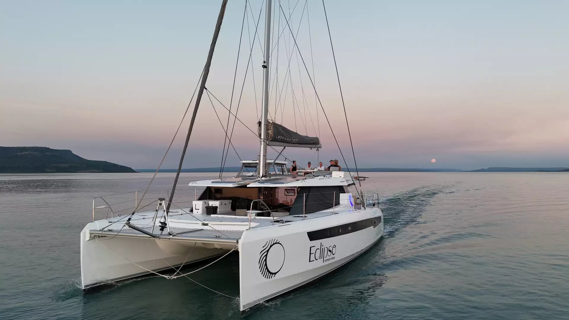 A low-angle shot from the water looking up at the back of the white catamaran Eclipse. Guests are visible on the top deck, with a full moon rising on the horizon under a soft pink and purple sky.