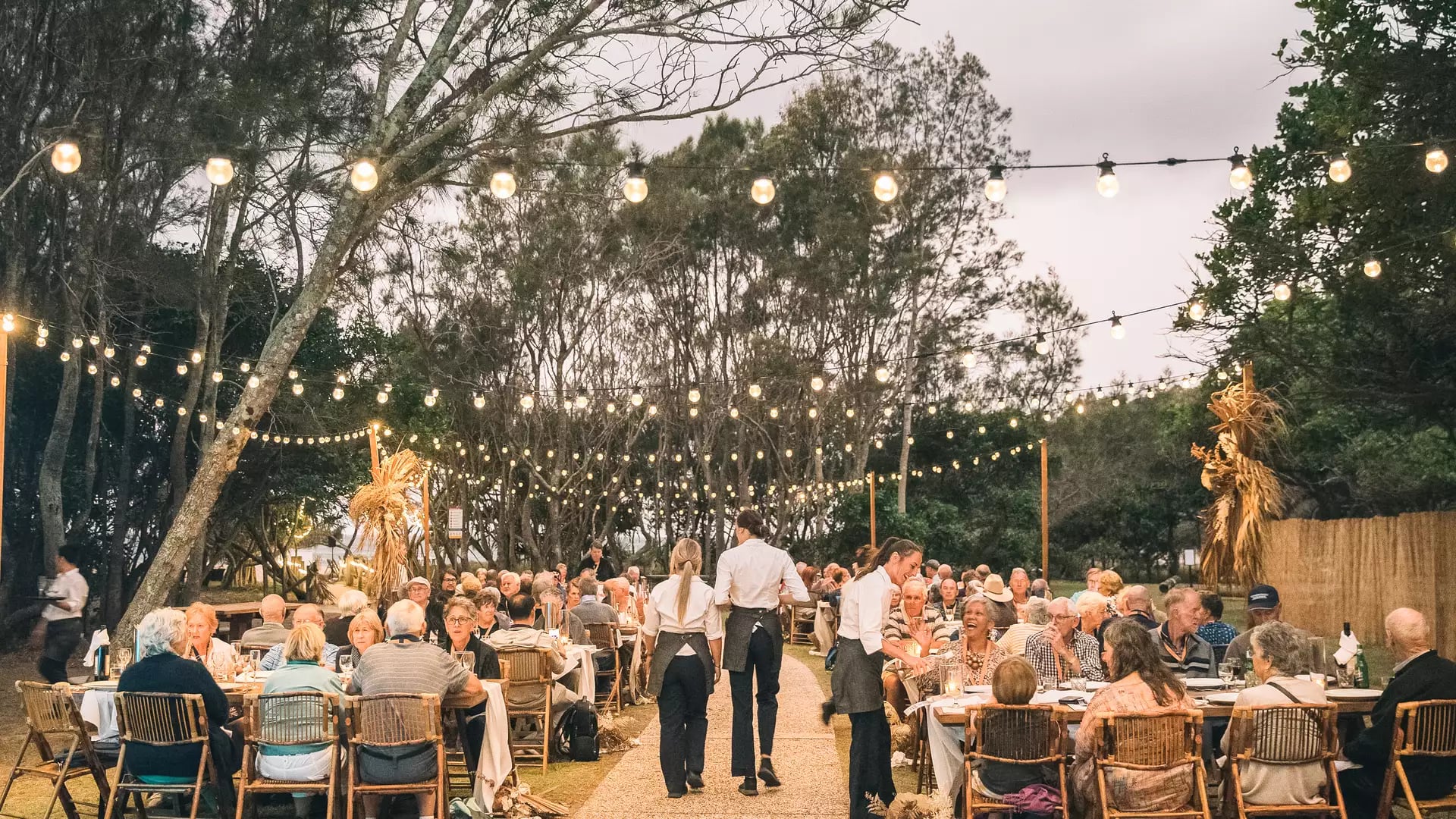 Waitstaff serving guests at an outdoor dinner party under festive string lights at dusk.