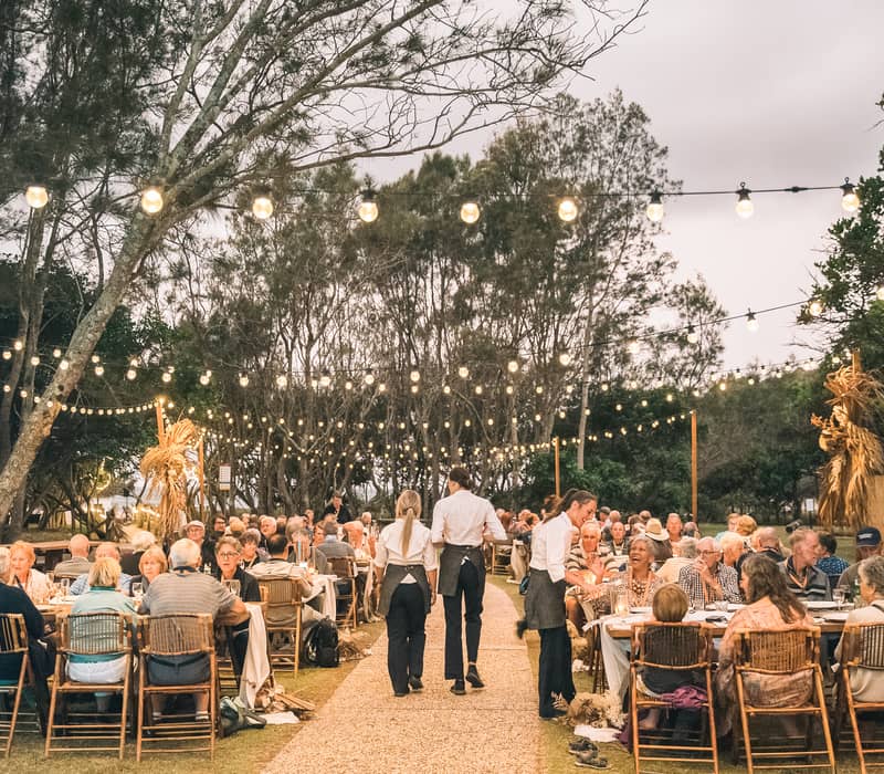 Waitstaff serving guests at an outdoor dinner party under festive string lights at dusk.