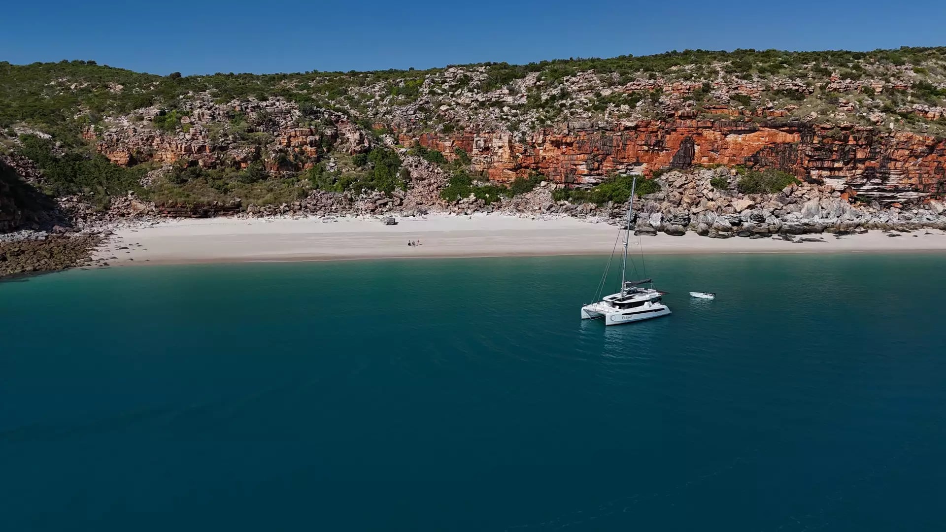 A wide aerial shot of a pristine white sand beach at the base of a tall, layered orange rock cliff. The Eclipse catamaran is anchored in the clear turquoise water just offshore.