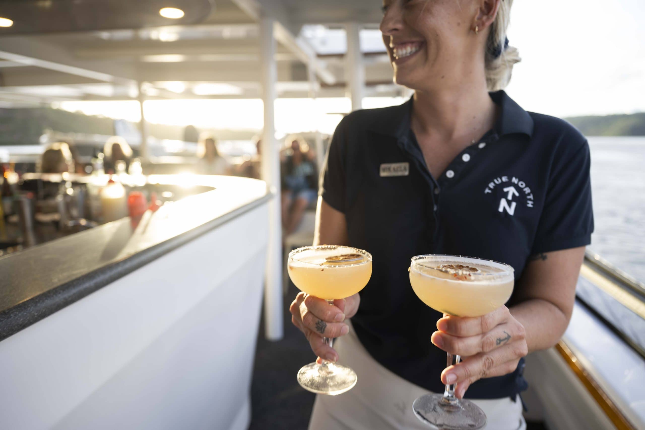 Close up of a smiling server holding two yellow cocktails in coupe glasses on a True North ship deck at sunset in the Kimberleys.