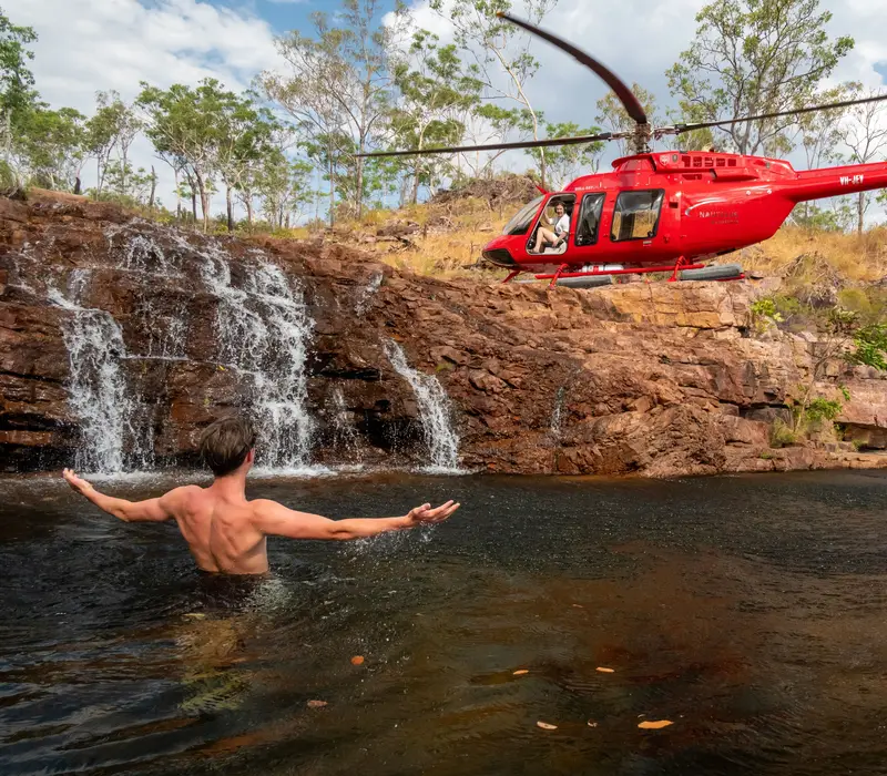 A red helicopter perched on rocks next to a person swimming in a natural waterfall pool in the Northern Territory.