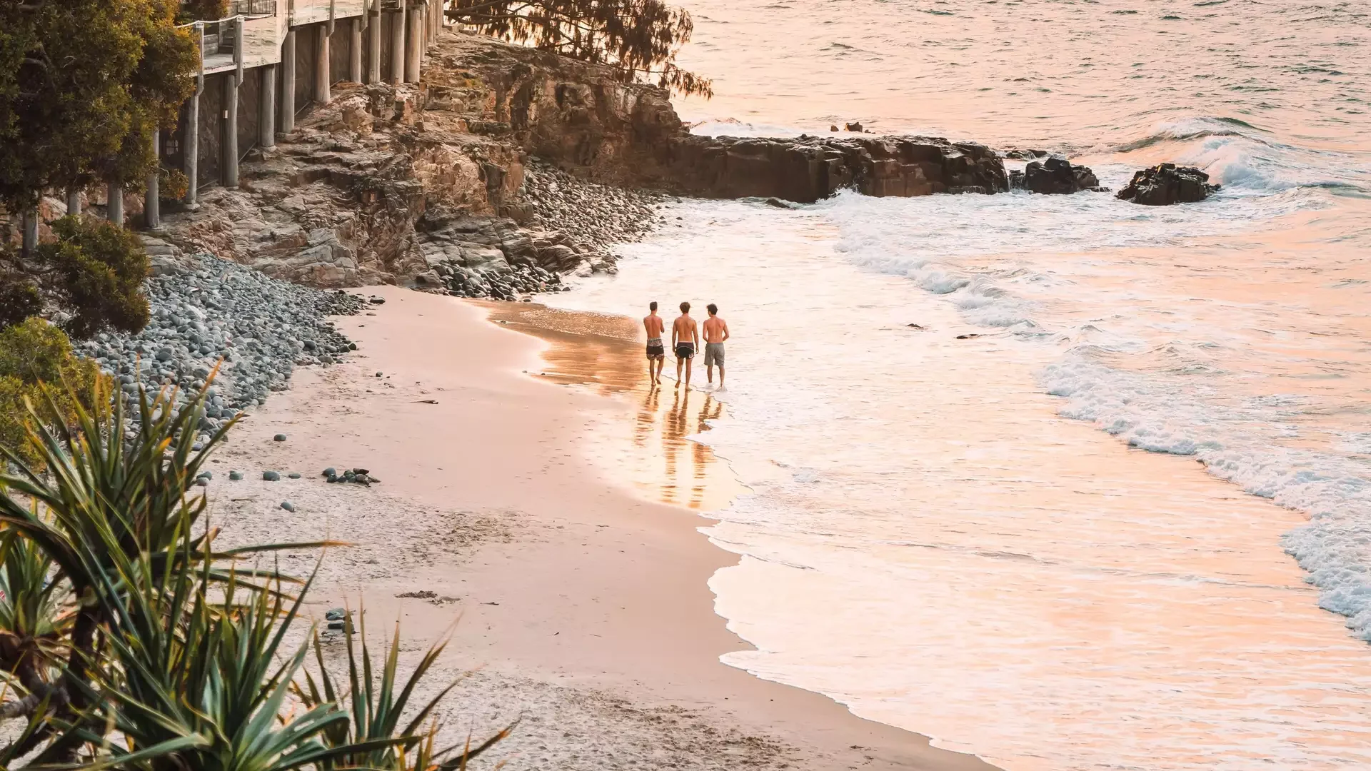 Three people walking on a beach at sunset with a glowing orange sky and mountains in the distance.