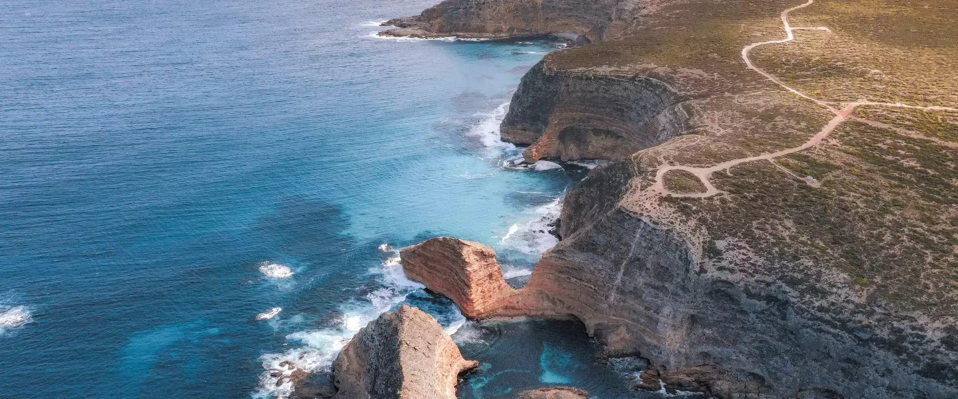 An expansive aerial shot captures the raw beauty of the Eyre Peninsula's Whalers Way, where high limestone cliffs meet the deep blue of the Southern Ocean. Dirt tracks wind along the cliff tops, offering spectacular viewpoints of the rugged sea stacks and surf below.